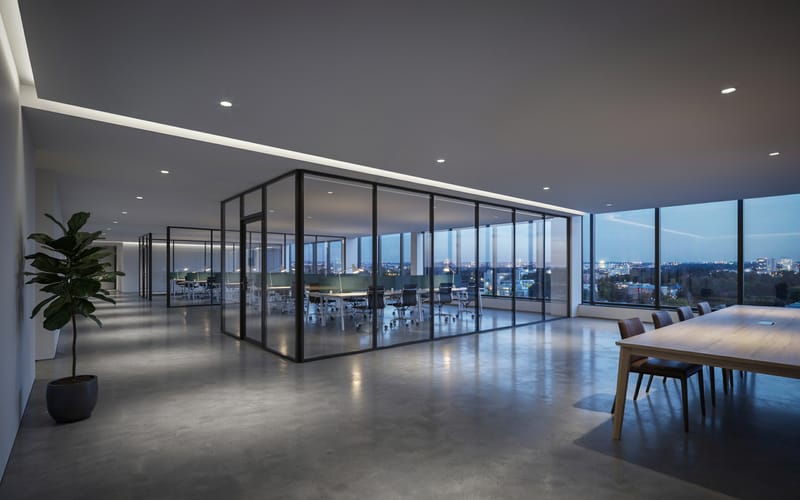A modern office corridor at dusk with a glass-walled meeting room and a city skyline through the windows